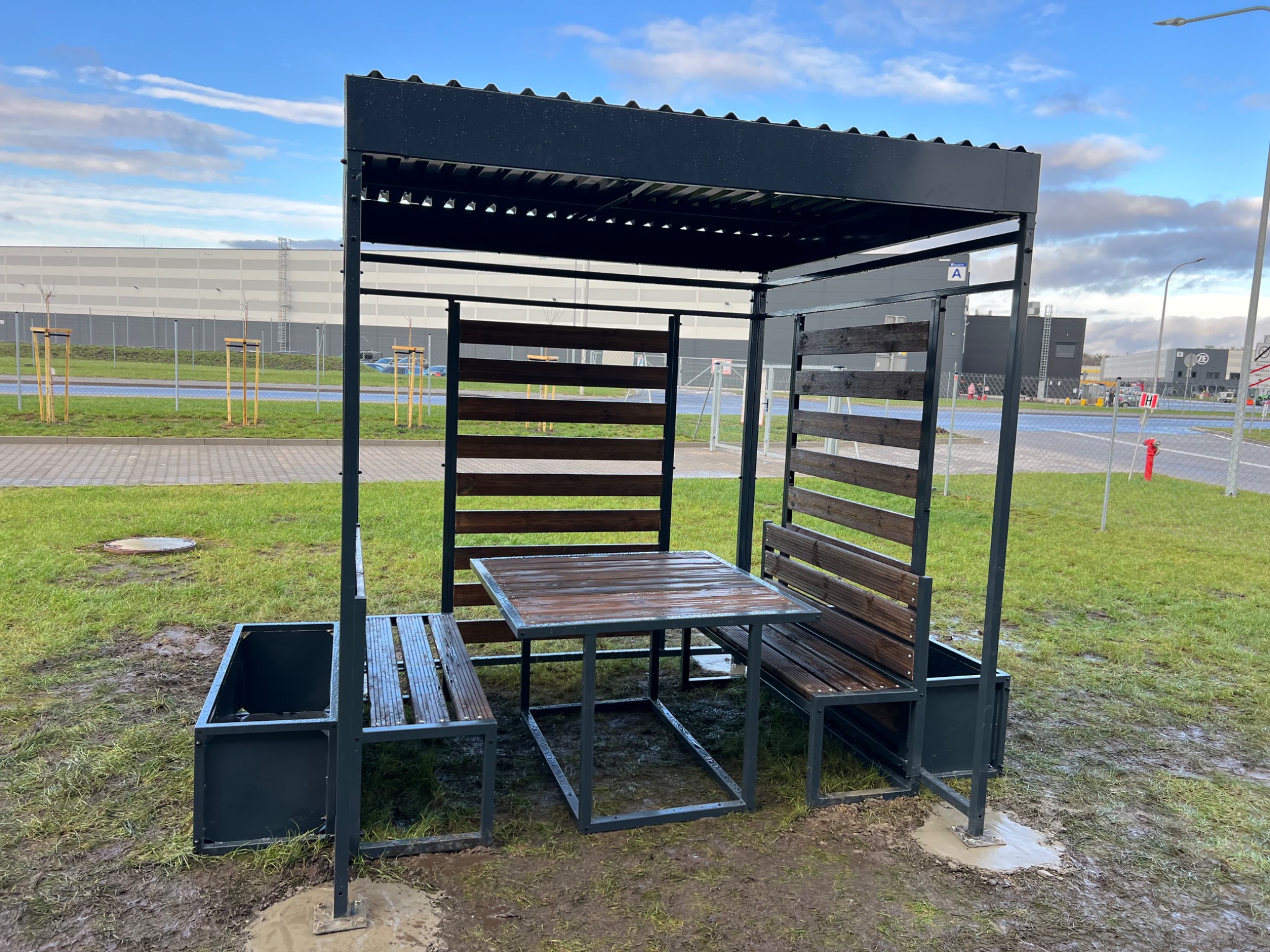 Metalowa altana z drewnianymi elementami, ławeczkami i stołem, umieszczona na mokrym trawniku w przemysłowej okolicy. Widoczne budynki magazynowe w tle.