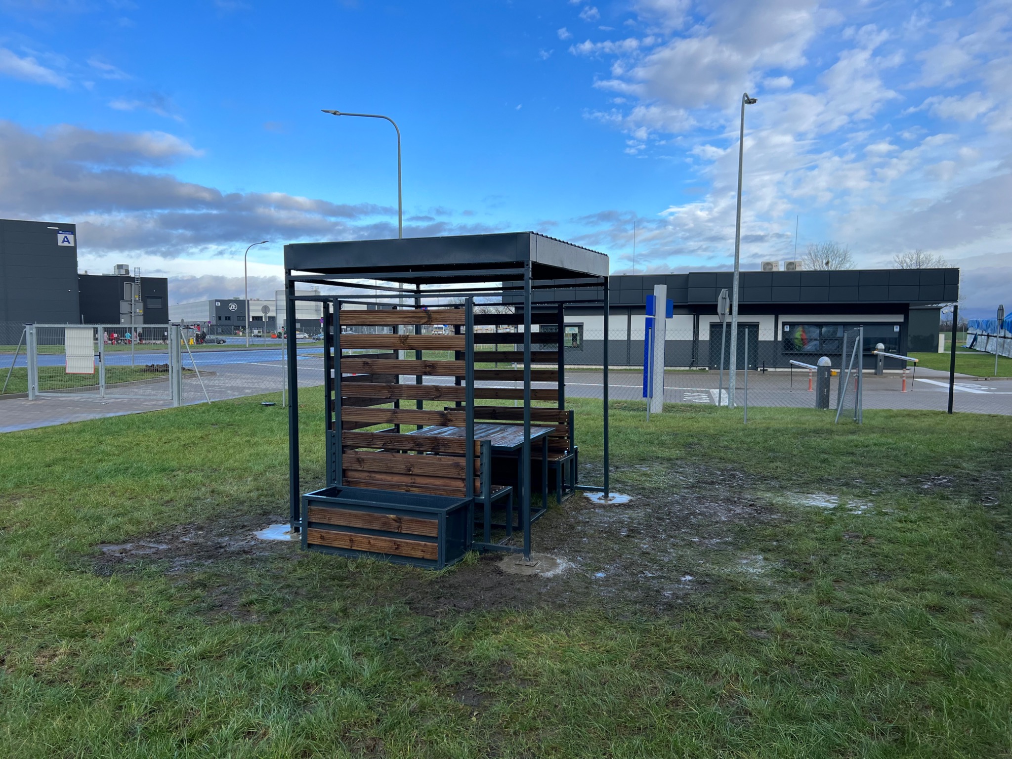 Zadaszona, metalowa konstrukcja z drewnianymi elementami, służąca jako miejsce do siedzenia i spożywania posiłków na zewnątrz, ustawiona na trawniku z widocznymi śladami po deszczu, w tle budynki...