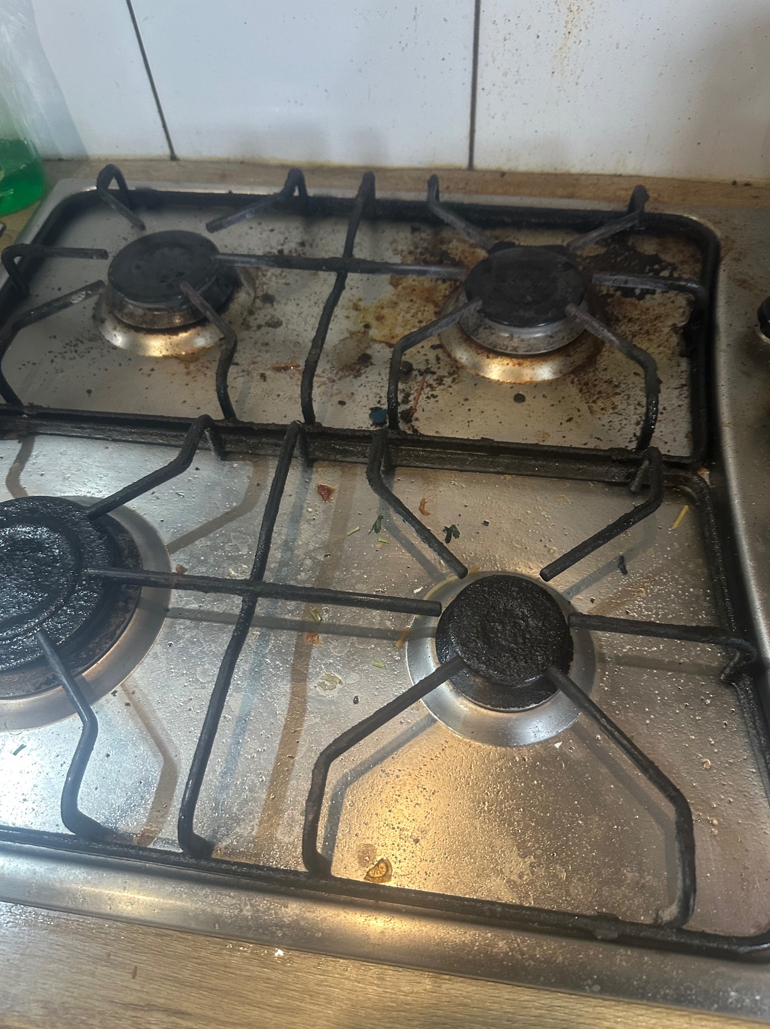 Zabrudzona, metalowa kuchenka gazowa z czterema palnikami, widoczne resztki jedzenia i przypalenia na powierzchni płyty.