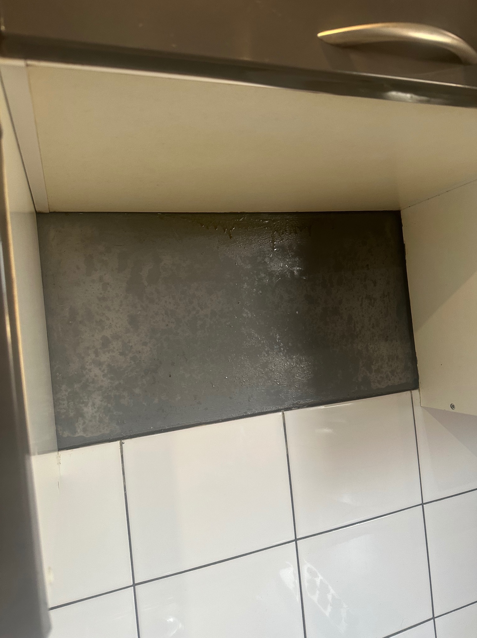 Wnętrze szafki kuchennej z widocznymi zabrudzeniami i plamami na dnie, poniżej białe płytki podłogowe, ujęcie z góry.