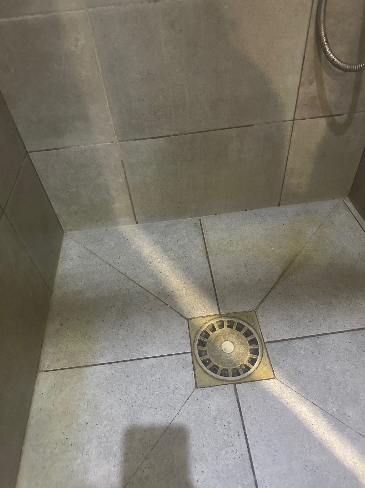 Szare płytki podłogowe w kabinie prysznicowej z mosiężnym odpływem liniowym, widoczne ślady użytkowania.
