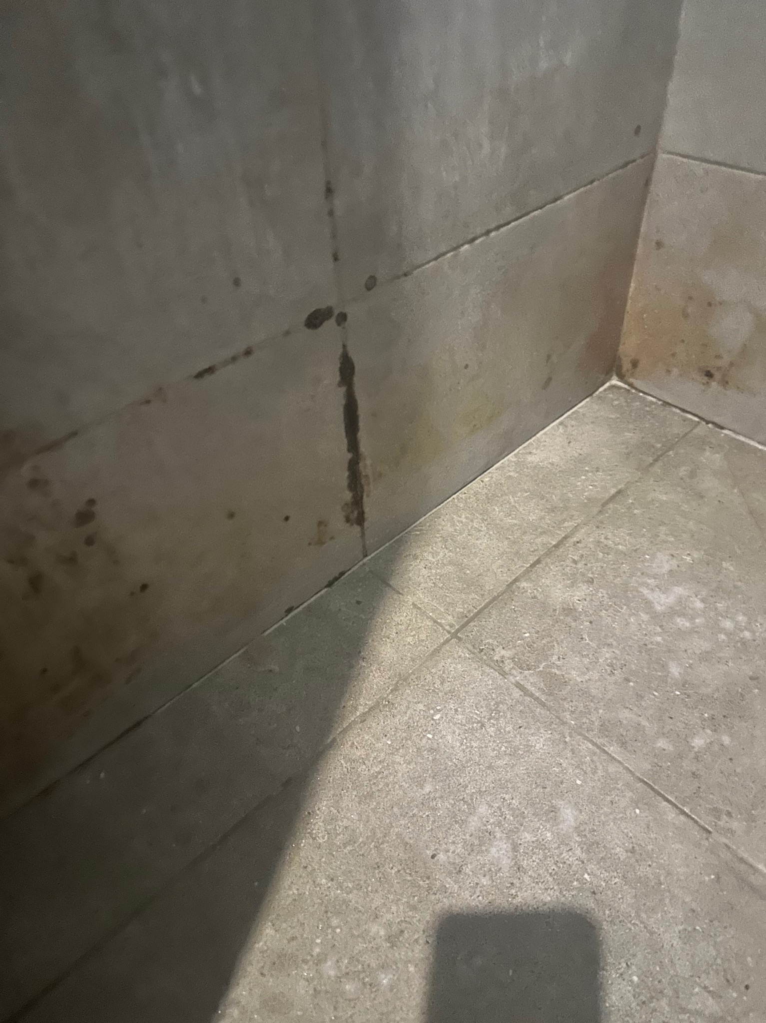 Szare płytki ścienne i podłogowe w rogu pomieszczenia z widocznymi plamami wilgoci i zaciekami, wskazujące na potrzebę sprzątania lub remontu.