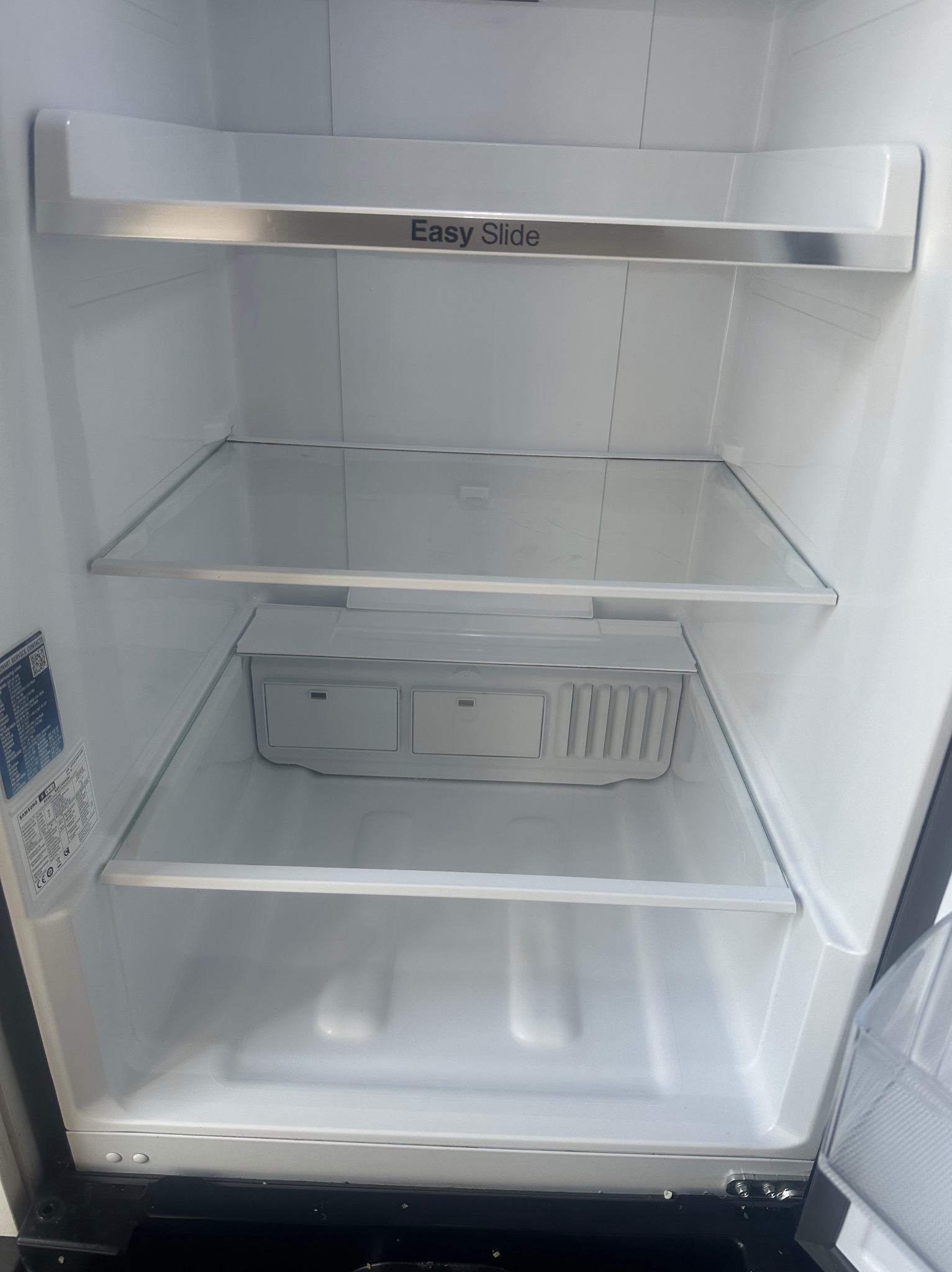 Pusta, czysta lodówka marki Samsung z półkami z hartowanego szkła i szufladą Easy Slide, gotowa do przechowywania żywności. Wnętrze oświetlone, bez zabrudzeń.