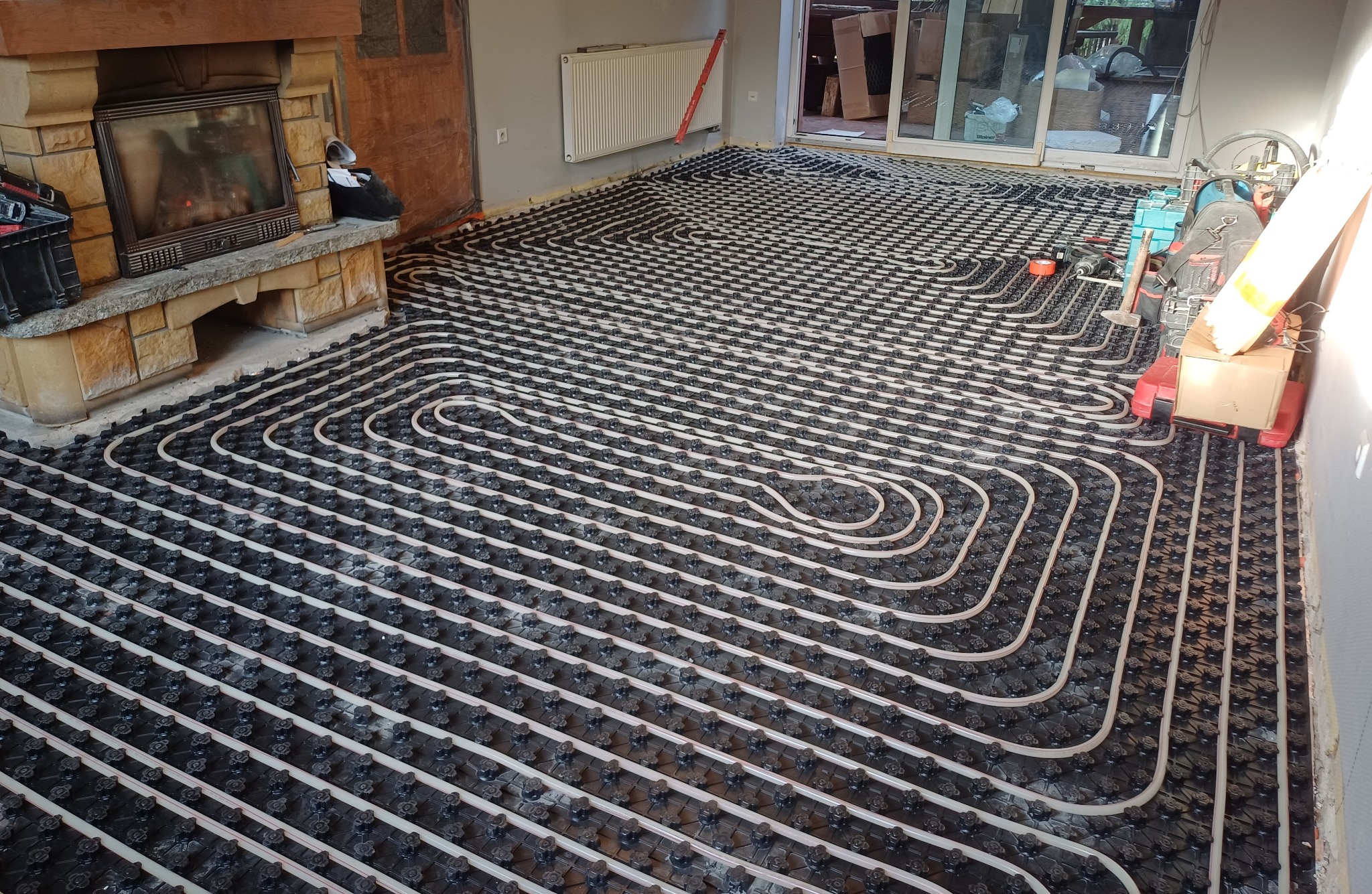 Rozłożona instalacja ogrzewania podłogowego z rurkami w kształcie meandrów, widoczny kominek w tle, narzędzia obok ściany, przygotowane do zalania wylewką.