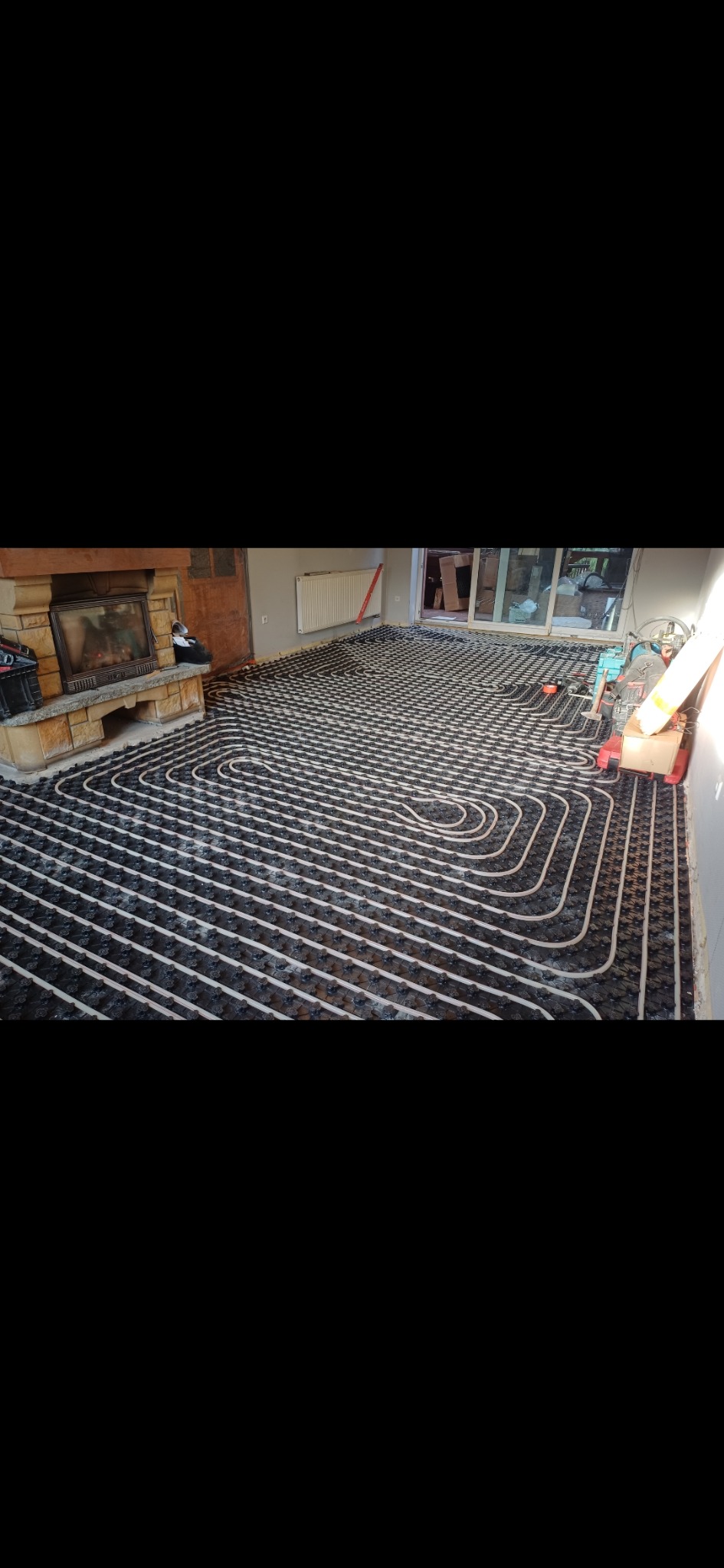 Rozłożone rury ogrzewania podłogowego na czarnej folii izolacyjnej w pomieszczeniu z kominkiem i grzejnikiem, widoczne narzędzia i materiały montażowe.