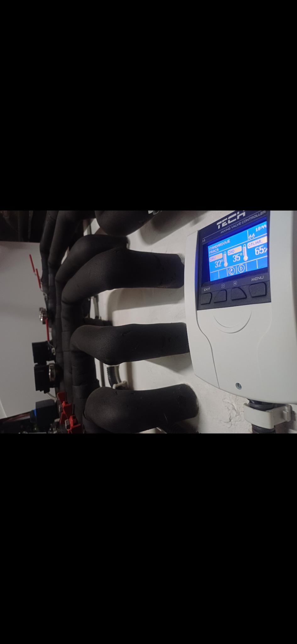 Regulator temperatury TECH z wyświetlaczem LCD, widoczne rury z izolacją termiczną, instalacja grzewcza w kotłowni.