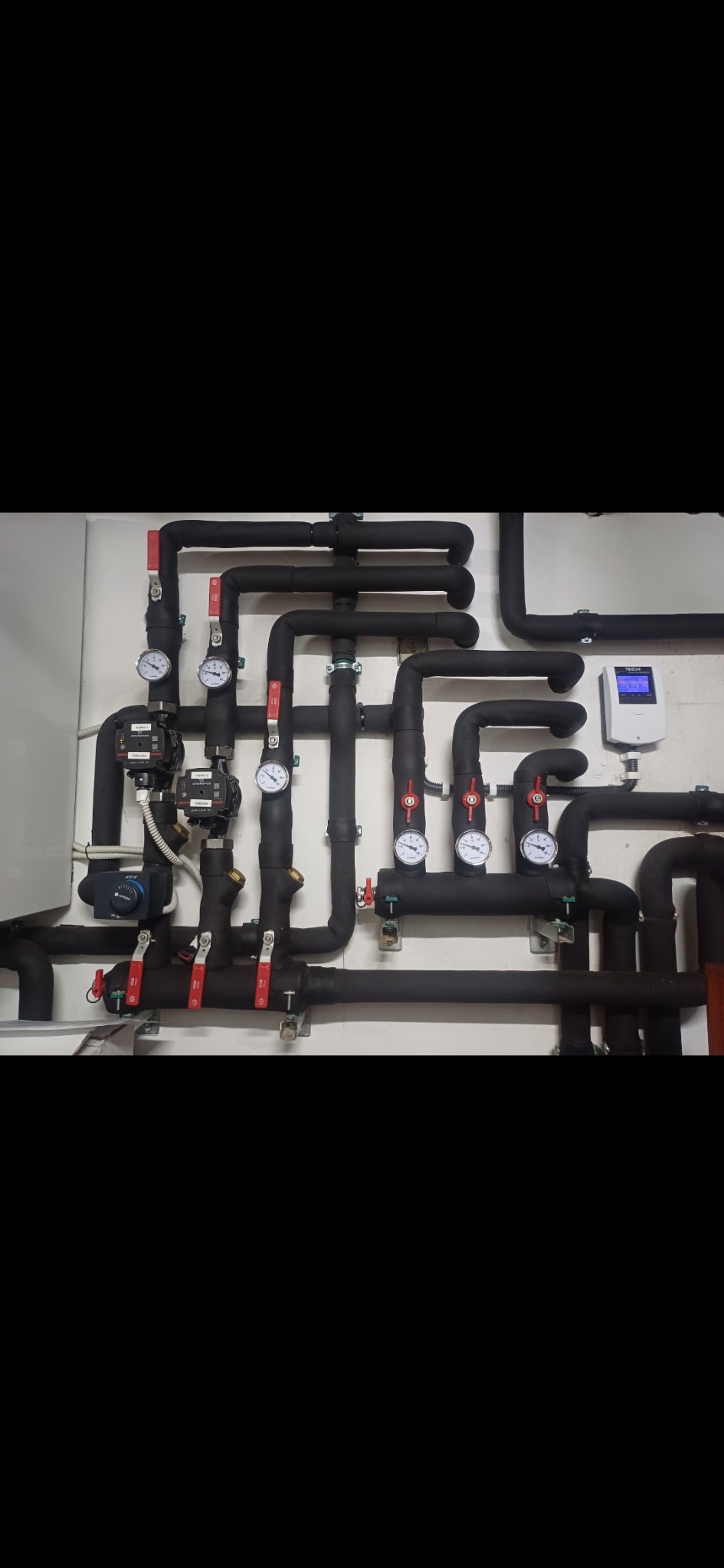 Czarny system rur z manometrami i zaworami zamontowany na białej ścianie, widoczny regulator temperatury.