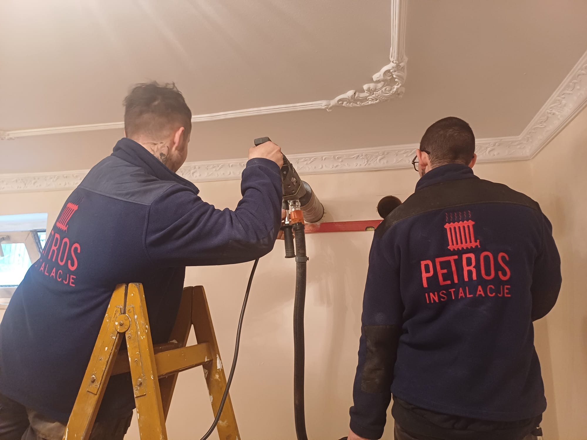 Dwóch instalatorów w granatowych bluzach z logo firmy wierci otwór w ścianie, używając wiertarki z odsysaniem pyłu. Jeden stoi na drewnianej drabinie, drugi przytrzymuje poziomnicę. Sufit zdobiony...