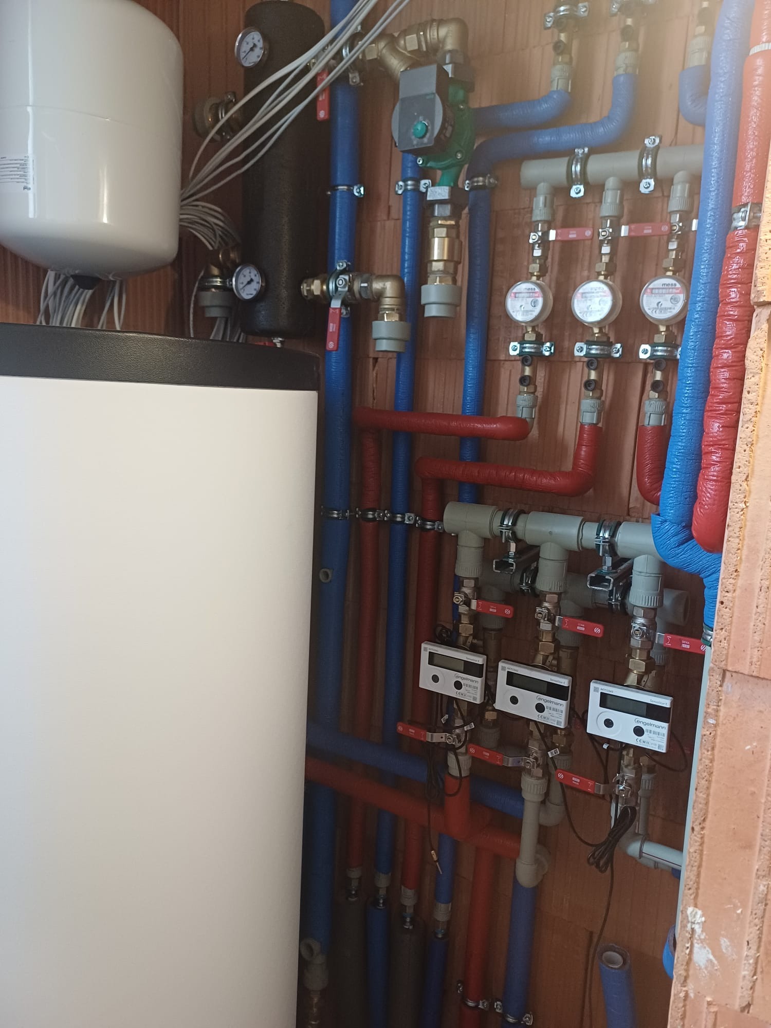 Rozbudowana instalacja centralnego ogrzewania z kotłem, zbiornikiem buforowym i rozdzielaczem z licznikami ciepła, zamontowana przy ścianie z cegły dziurawki.