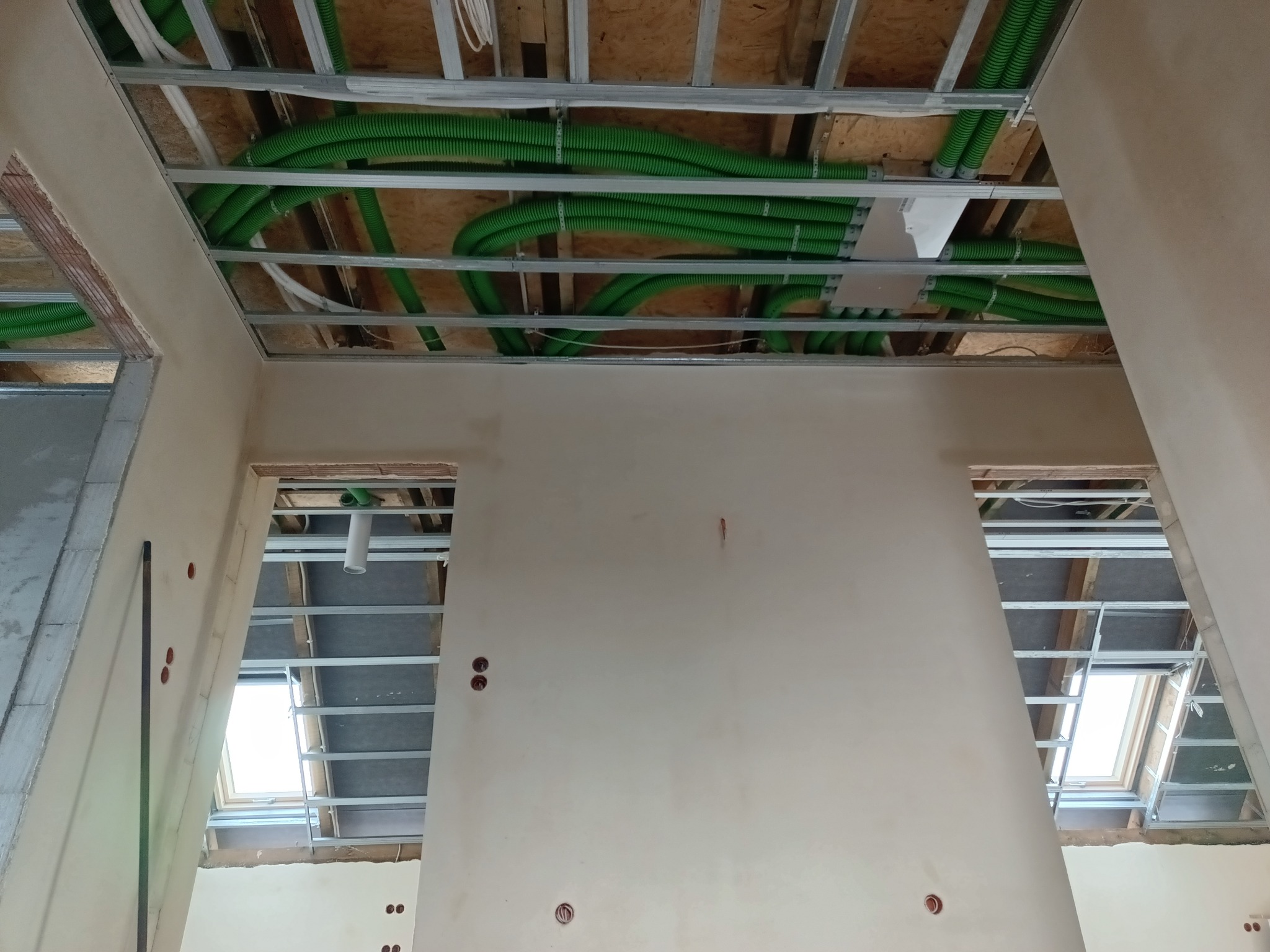 Zielone rury instalacyjne ułożone pod sufitem podwieszanym w surowym wnętrzu. Widoczne okna dachowe i przygotowane punkty elektryczne w ścianach.