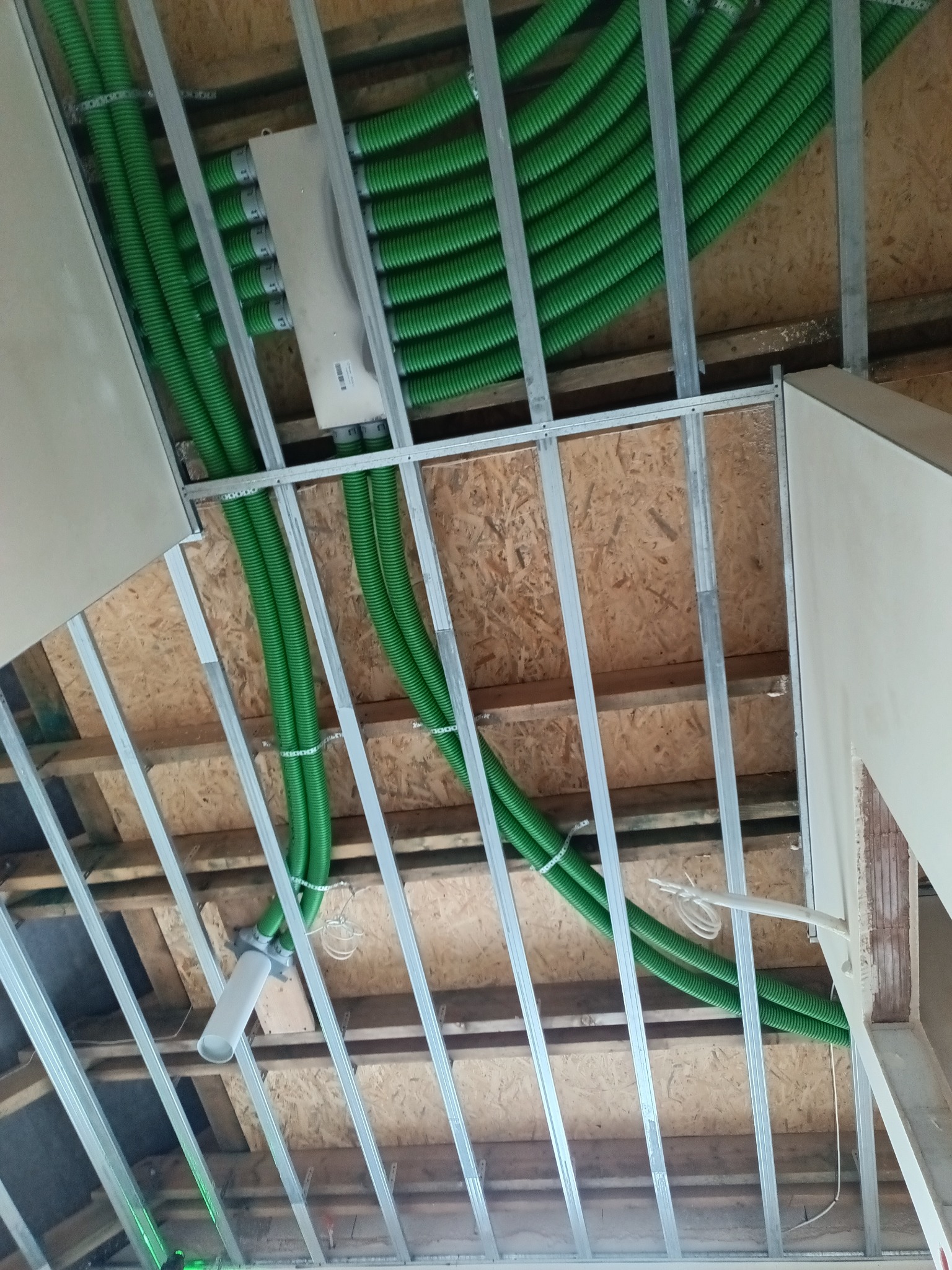 Zielone rury wentylacyjne ułożone pod stropem z drewnianych belek i metalowych profili. Widoczna skrzynka rozdzielcza i fragment ściany. Ujęcie z dołu.