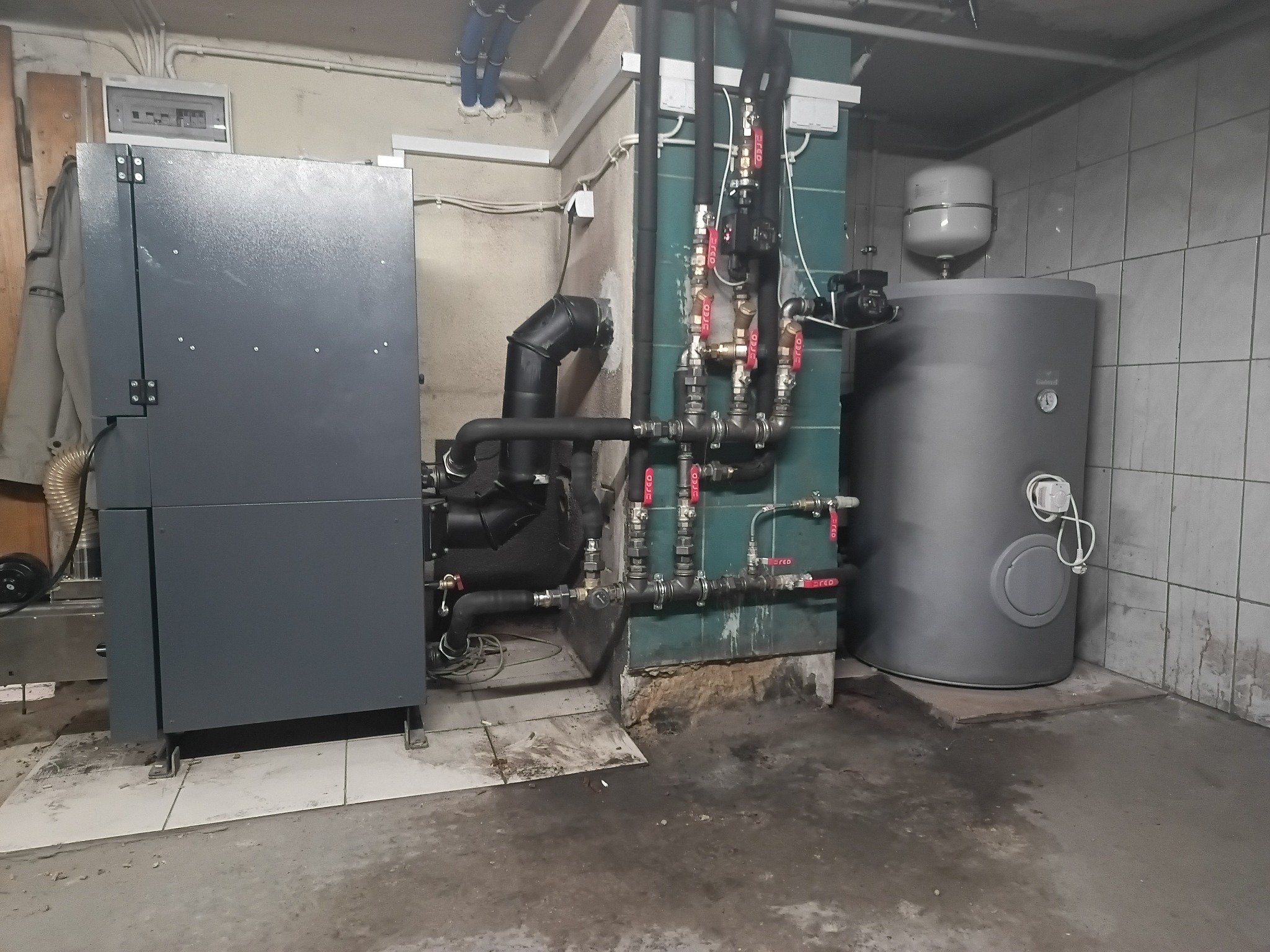 Szary piec CO z licznymi rurami i zaworami w kotłowni z wykafelkowanymi ścianami. Widoczny zbiornik wyrównawczy i instalacja.