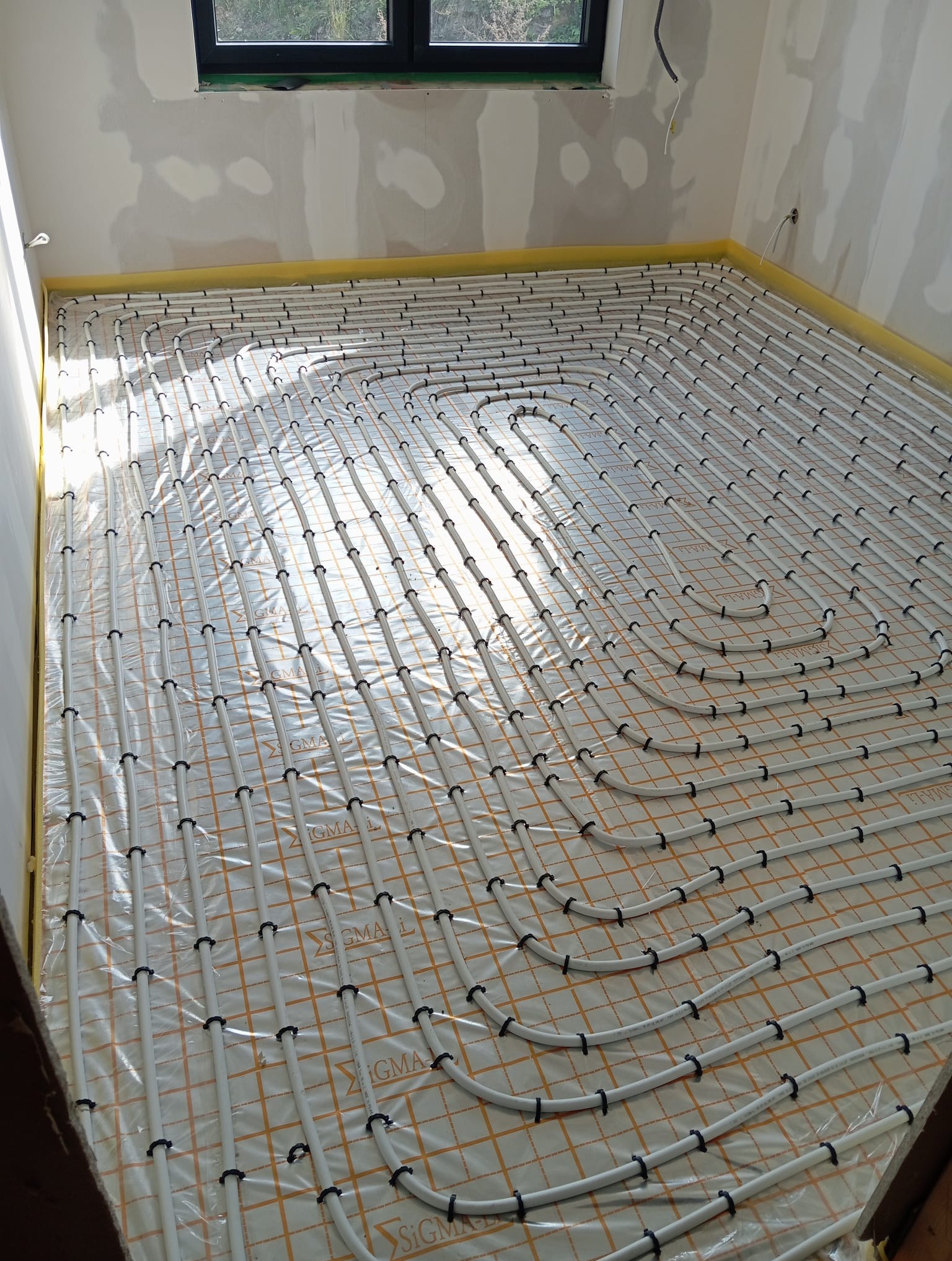 Białe rury ogrzewania podłogowego ułożone na folii z siatką, przymocowane czarnymi uchwytami w jasnym pomieszczeniu w trakcie instalacji. Widok z góry, surowy stan ścian.