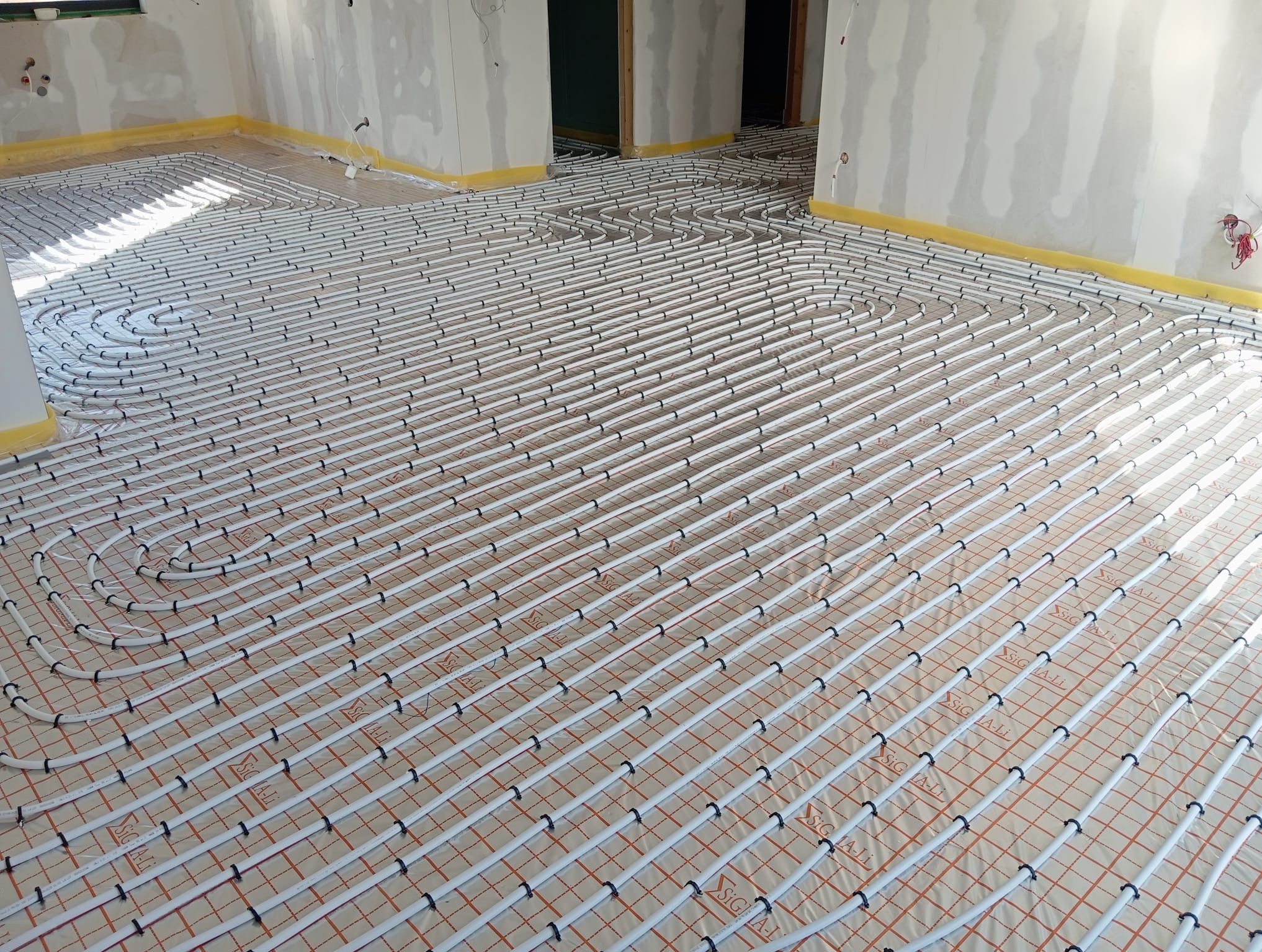 Białe rury ogrzewania podłogowego ułożone na folii w Pszczynie, widok z góry. Instalacja przed wylaniem posadzki, żółta taśma na ścianach.