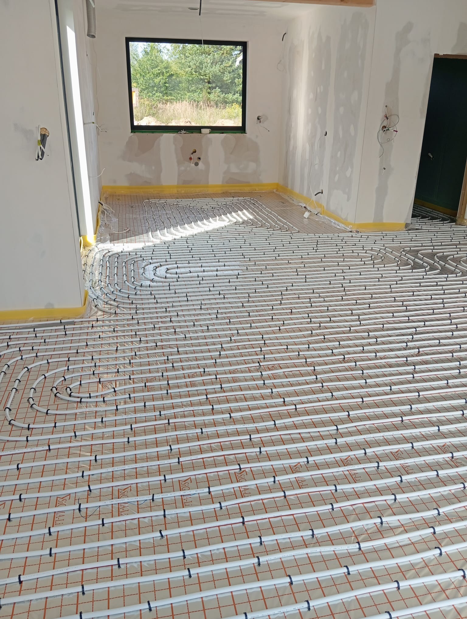 Białe rury ogrzewania podłogowego ułożone na pomarańczowej macie w surowym wnętrzu z oknem. Żółta taśma zabezpiecza ściany. Widoczne niedokończone instalacje.