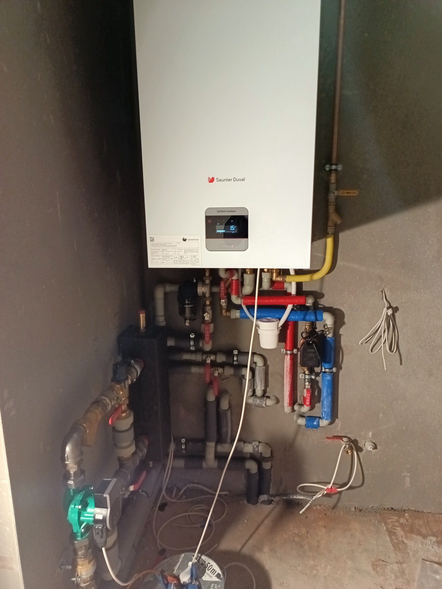 Biały piec gazowy Saunier Duval zamontowany na szarej ścianie, z widoczną instalacją hydrauliczną z rur w kolorach czerwonym, niebieskim i żółtym. Widoczne liczne zawory i złączki.
