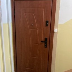 Fach-bud Paweł Piechal - Brązowe drzwi wejściowe z czarną klamką i zamkami, zamontowane w korytarzu z żółtymi ścianami. Widoczne detale wykończenia.