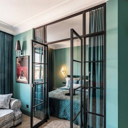 Fach-bud Paweł Piechal - Eleganckie, szklane drzwi dwuskrzydłowe z czarną ramą oddzielają pokój. W tle widoczne łóżko i zasłony w odcieniach zieleni i błękitu. Parkiet w geometryczny wzór.