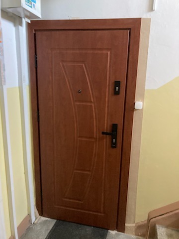 Brązowe drzwi wejściowe z czarną klamką i zamkami, zamontowane w korytarzu z żółtymi ścianami. Widoczne detale wykończenia.