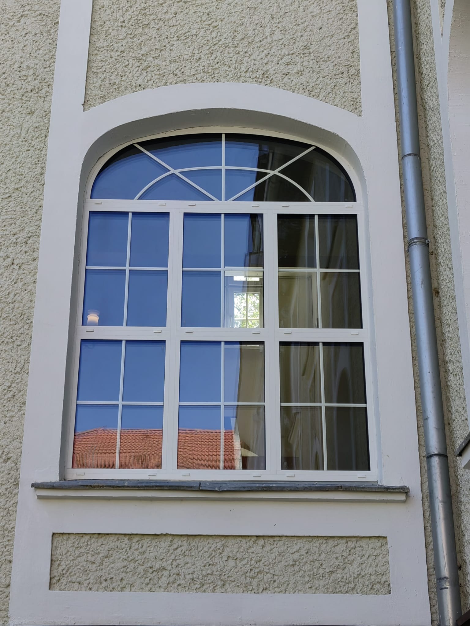 Białe, łukowe okno z kratownicą na teksturowanej, kremowej elewacji budynku. Widoczny fragment rynny po prawej stronie. W szybach odbija się czerwony dach.