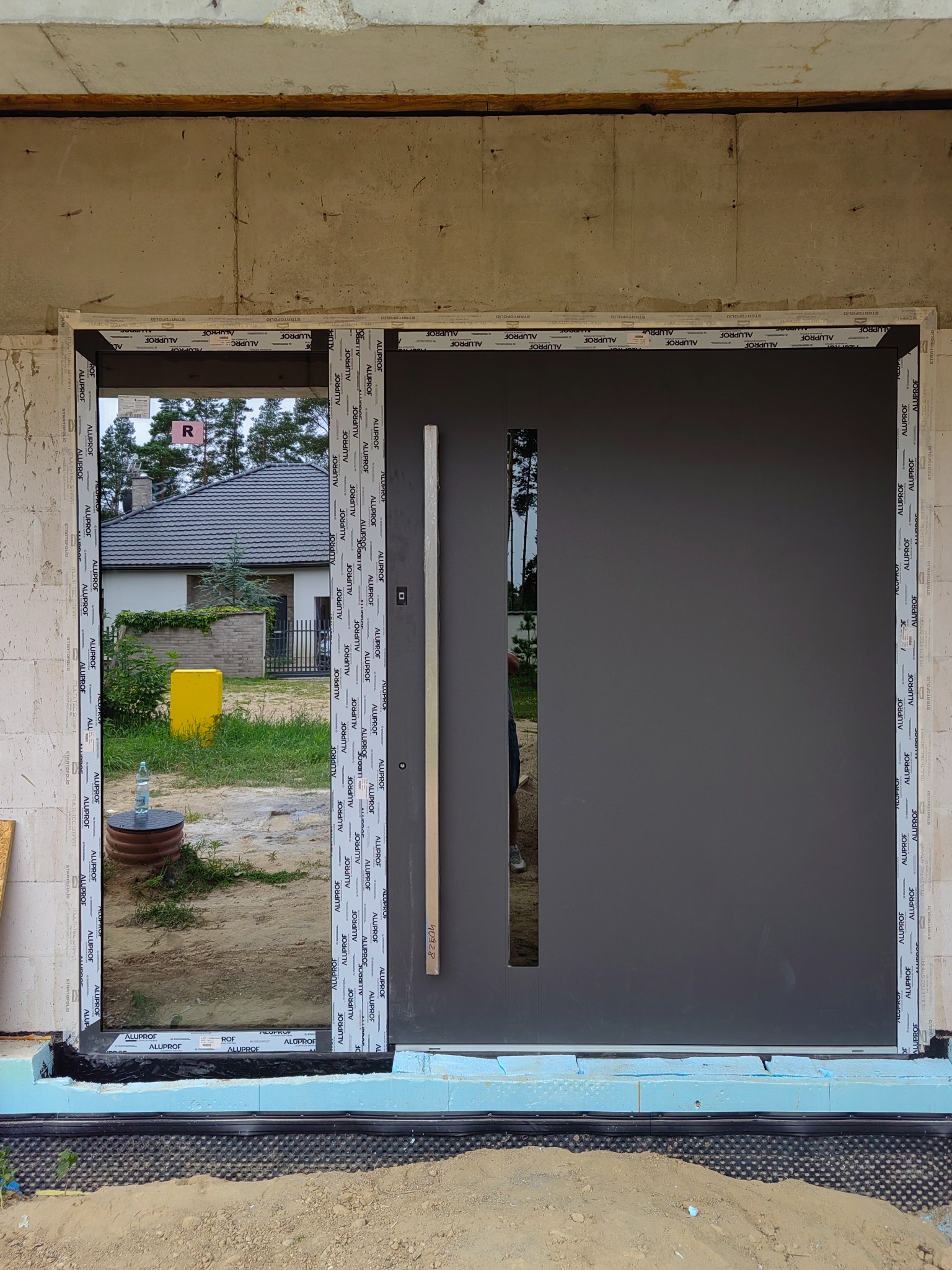 Nowe, grafitowe drzwi wejściowe z przeszkleniem bocznym, zamontowane w surowym stanie budynku. Widoczne materiały izolacyjne i otoczenie budowy.