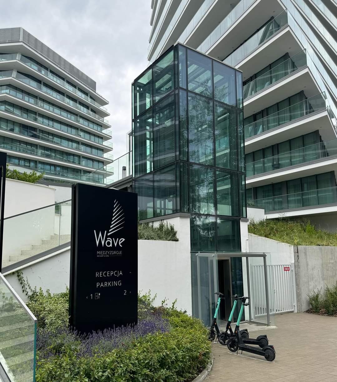 Nowoczesny budynek z przeszkloną windą zewnętrzną i tablicą informacyjną 'Wave'. Dwa elektryczne skutery zaparkowane przed wejściem. Architektura minimalistyczna, zieleń.