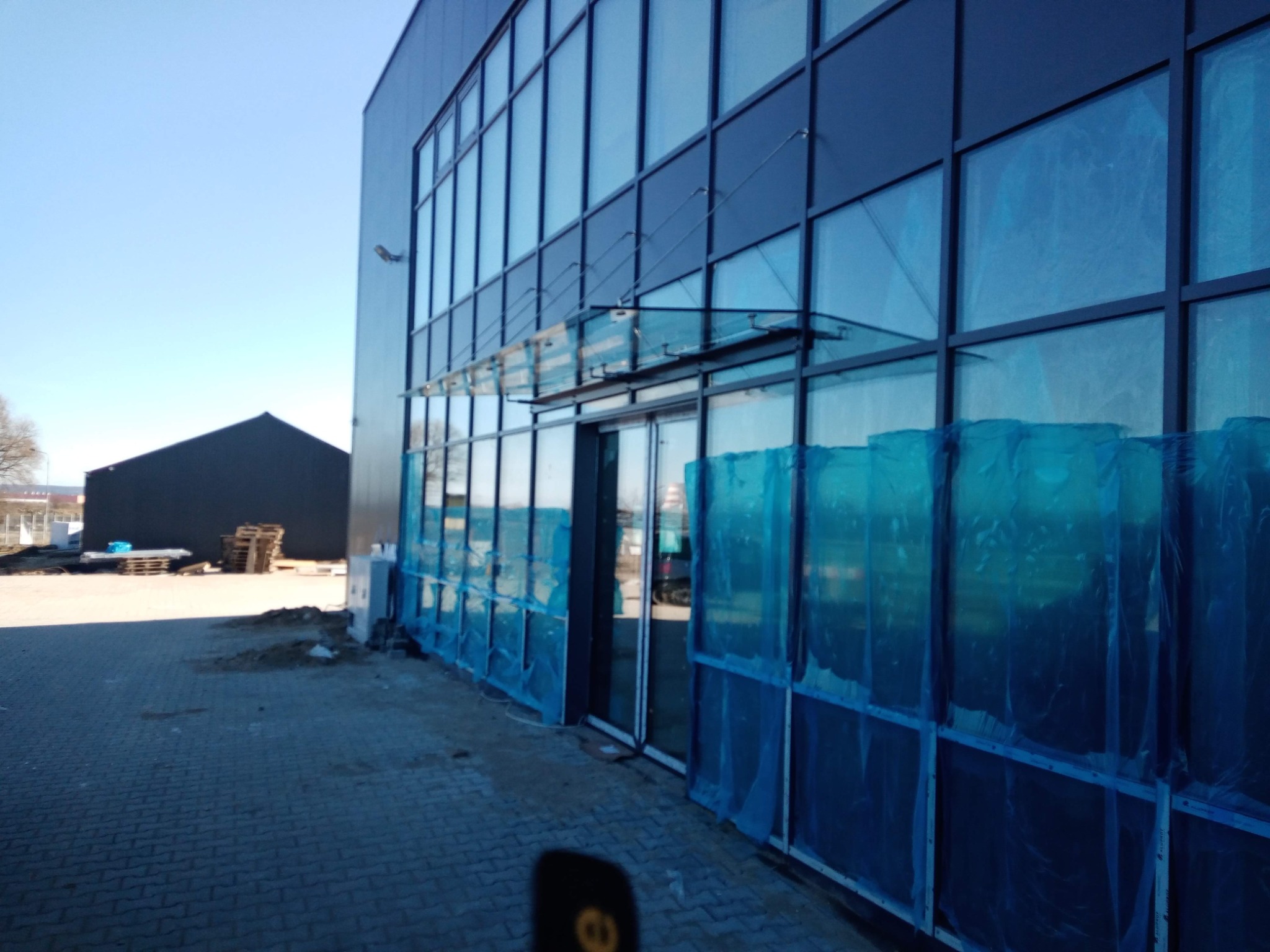 Nowoczesny budynek biurowy z oszkloną fasadą i szklanym zadaszeniem nad wejściem, zabezpieczony folią ochronną w trakcie prac wykończeniowych.