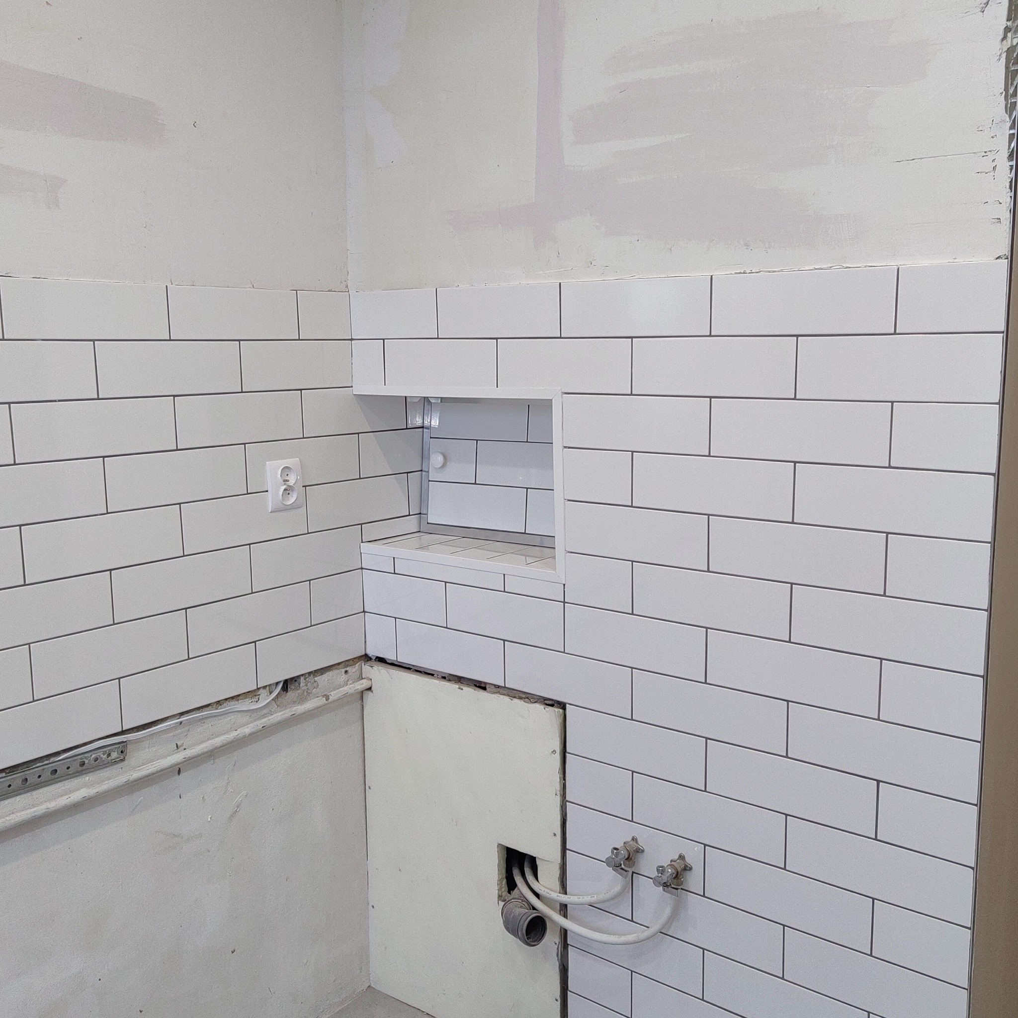 Białe, prostokątne płytki ułożone na ścianie łazienki, z wnęką na kosmetyki i widoczną instalacją wodną. Częściowo wykończona ściana.