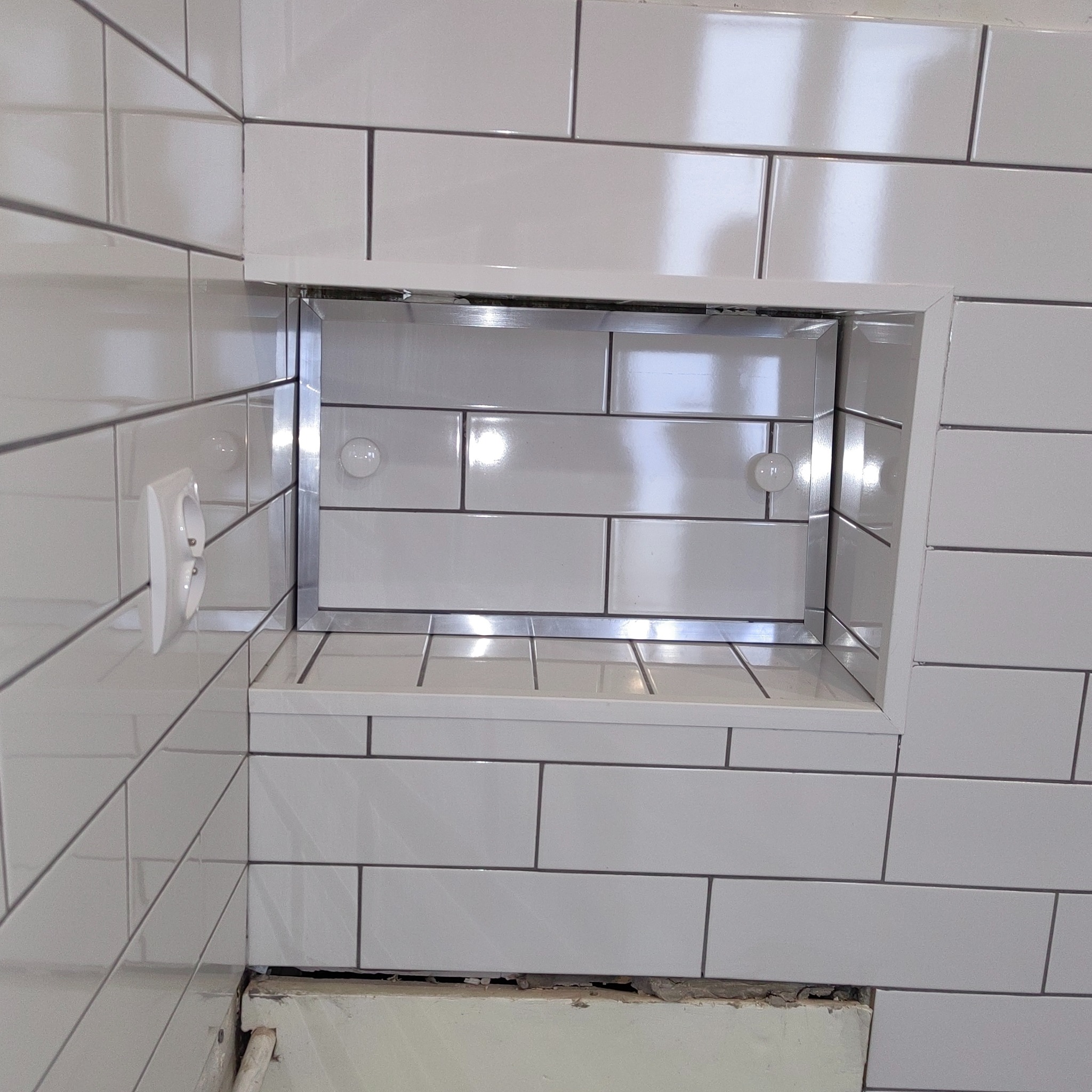 Białe, błyszczące płytki w łazience z wnęką w ścianie wyłożoną lustrzanymi płytkami. Widoczne gniazdko elektryczne. Minimalistyczny design.