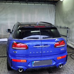 Myjnia samochodowa ZUBART - Niebieski Mini Clubman JCW po umyciu, widziany z tyłu, z widocznymi kroplami wody na karoserii i mokrą kostką brukową pod nim, wewnątrz zadaszonej myjni.
