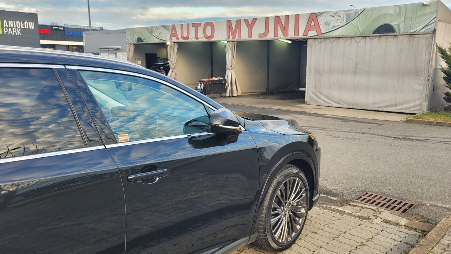 Częściowy widok czarnego samochodu na tle myjni samochodowej w Częstochowie, z widocznym napisem 'AUTO MYJNIA' nad stanowiskami.