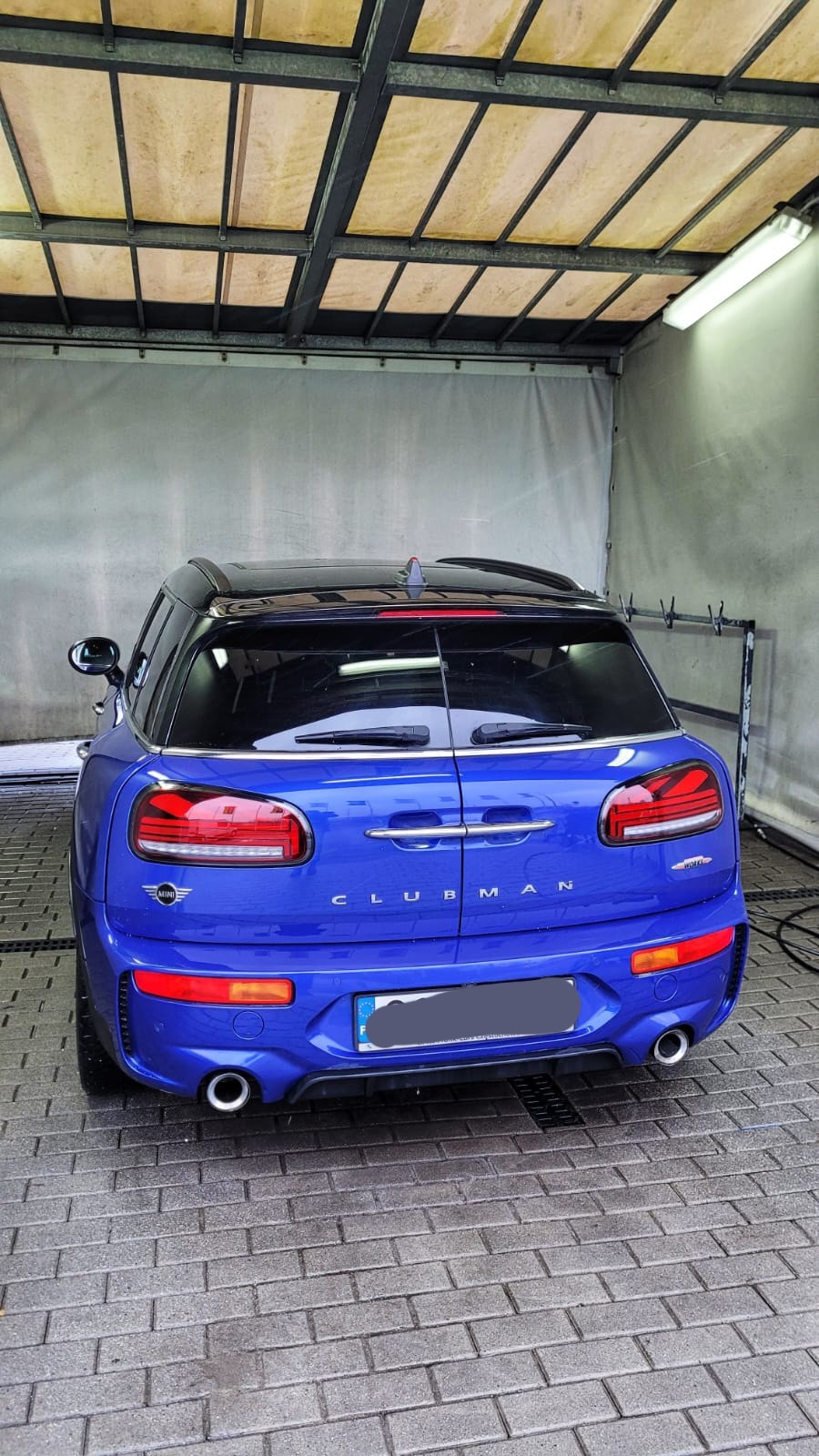 Niebieski Mini Clubman JCW po umyciu, widziany z tyłu, z widocznymi kroplami wody na karoserii i mokrą kostką brukową pod nim, wewnątrz zadaszonej myjni.