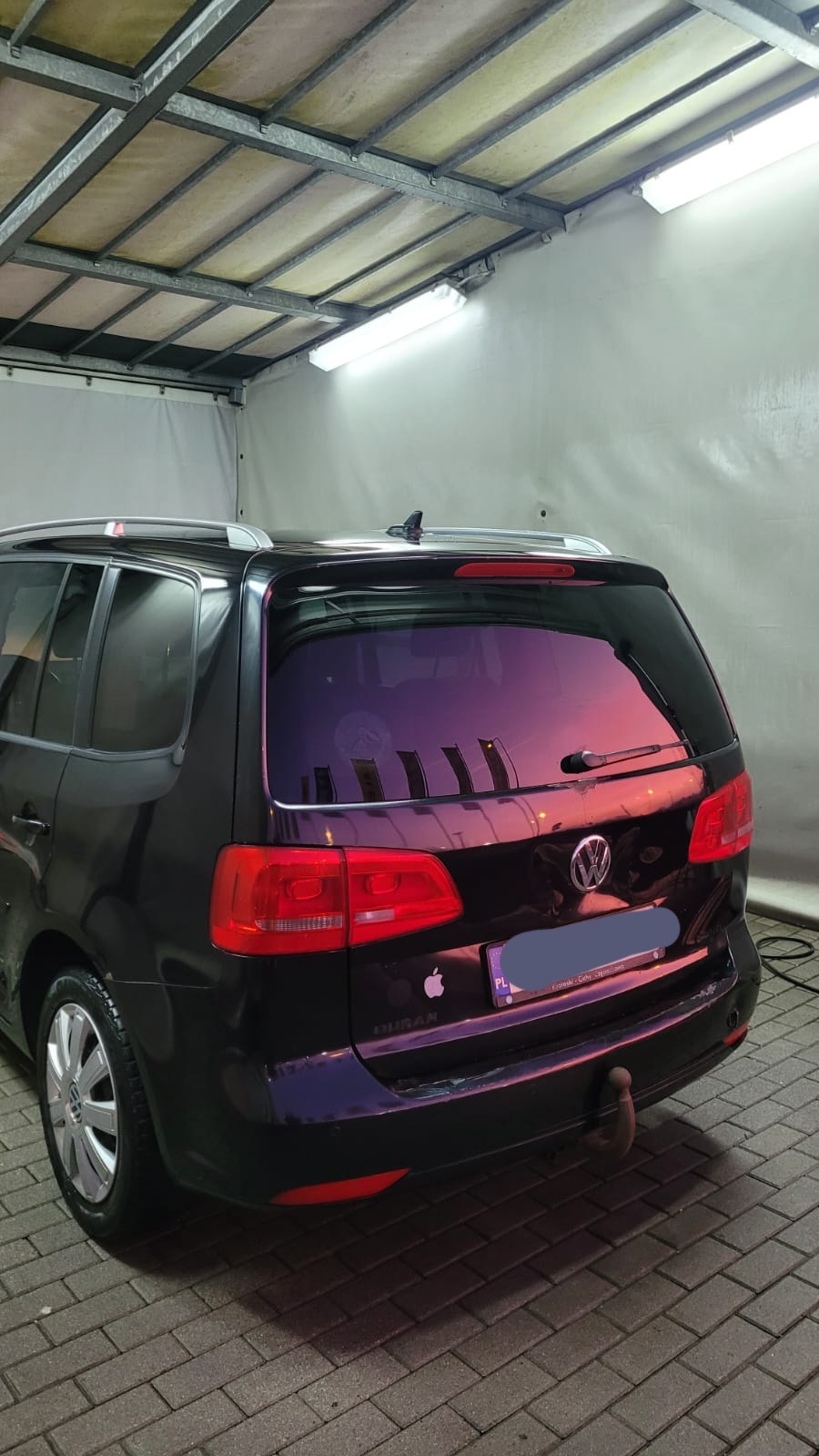 Czarny Volkswagen Sharan po umyciu, widoczny tył pojazdu z mokrą karoserią, na kostce brukowej, w oświetlonym pomieszczeniu myjni.