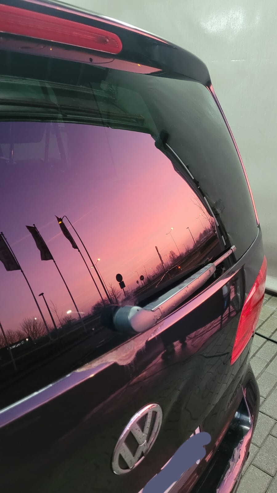 Odbicie różowego nieba z flagami i latarniami w brudnej szybie tylnej klapy czarnego samochodu Volkswagen.