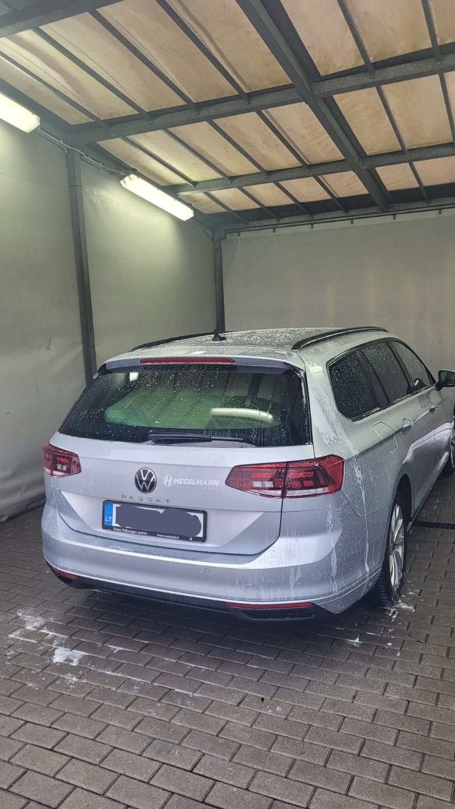 Srebrny samochód kombi marki Volkswagen Passat, z mokrą karoserią w trakcie mycia w zadaszonej myjni, widok z tyłu, ceglana posadzka.