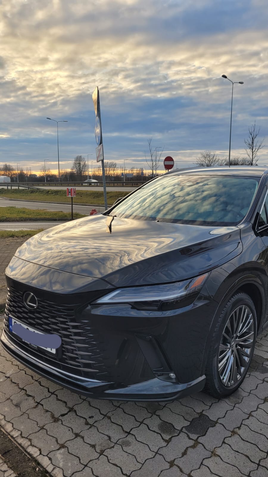 Czarny samochód Lexus RX, widok z przodu na tle pochmurnego nieba o zachodzie słońca, odbijającego się w przedniej szybie i karoserii.