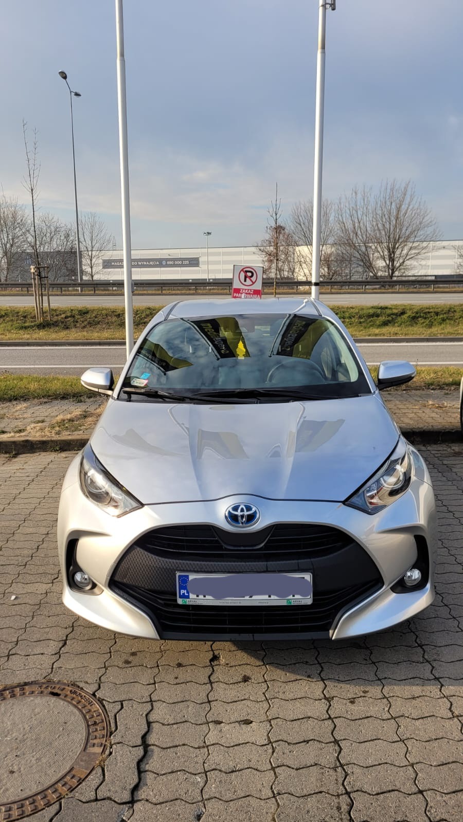 Srebrny Toyota Yaris zaparkowany na brukowanym parkingu, widok z przodu, z widocznym znakiem zakazu parkowania i budynkiem w tle.