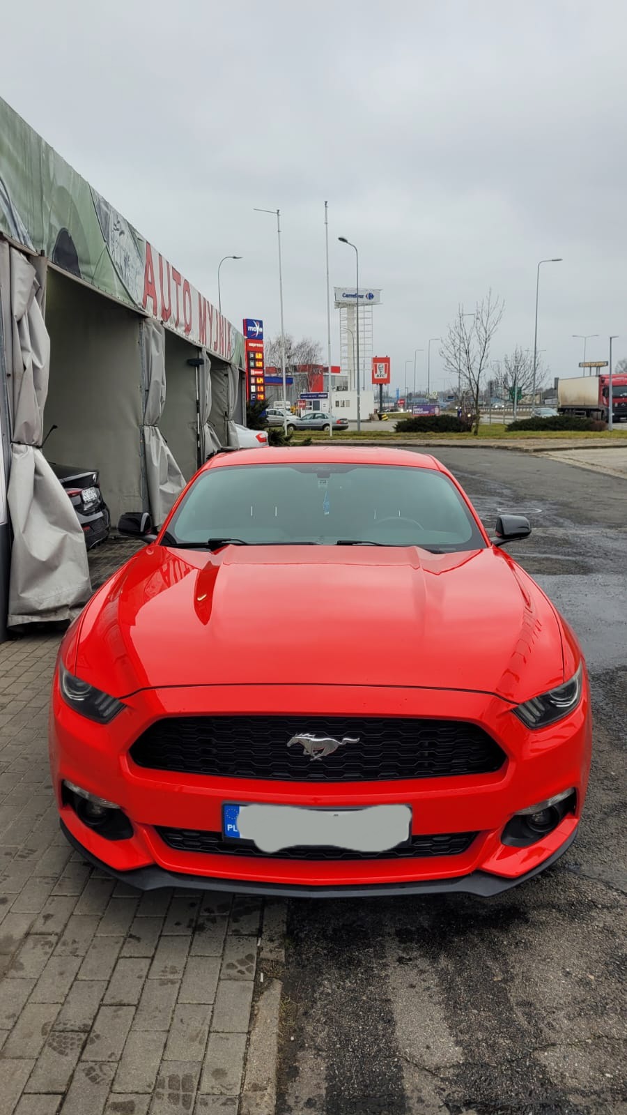 Czerwony Ford Mustang przed wjazdem do auto myjni z widoczną tablicą z cenami paliw w tle.