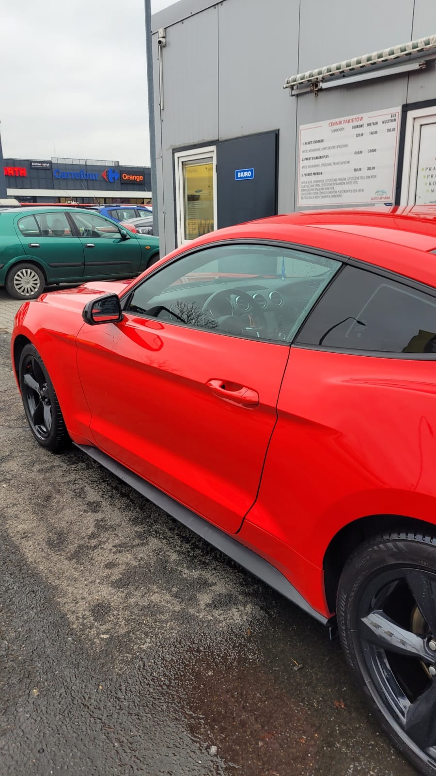 Czerwony Ford Mustang na parkingu myjni samochodowej, widoczny cennik pakietów mycia, mokry asfalt.