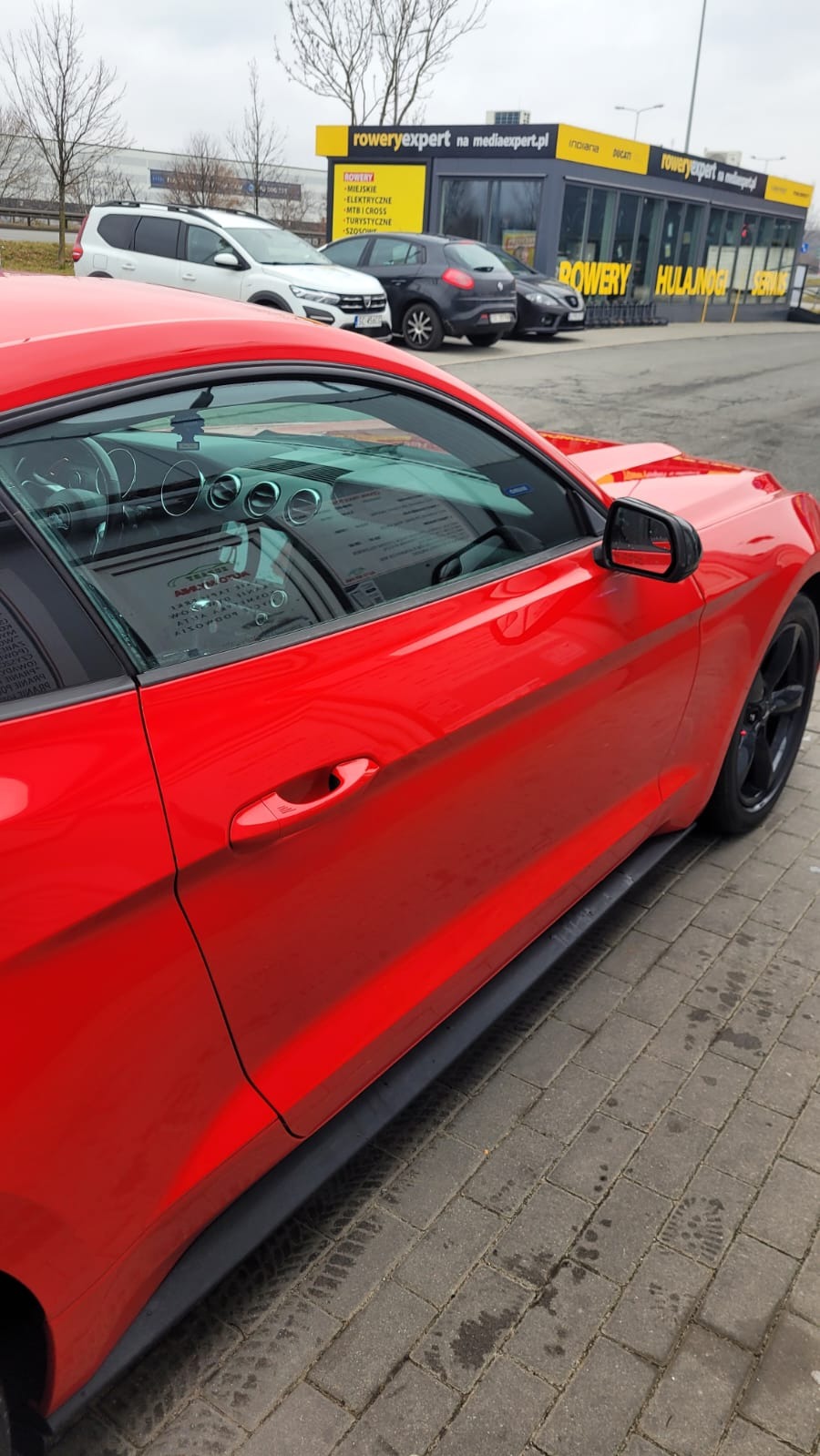 Czerwony samochód osobowy zaparkowany na kostce brukowej, widoczne odbicie w szybie i salon rowerowy w tle.