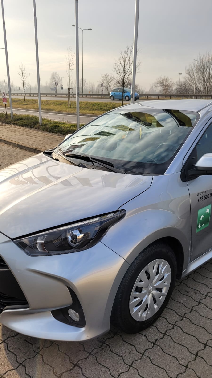 Srebrny samochód osobowy z naklejką z numerem telefonu i logo firmy na drzwiach, zaparkowany na kostce brukowej, w tle droga i zamglone drzewa.