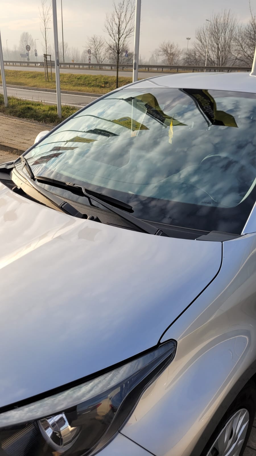 Srebrny samochód z widocznymi wycieraczkami i odbiciem chmur na przedniej szybie, z zawieszką zapachową i odblaskami na lakierze.