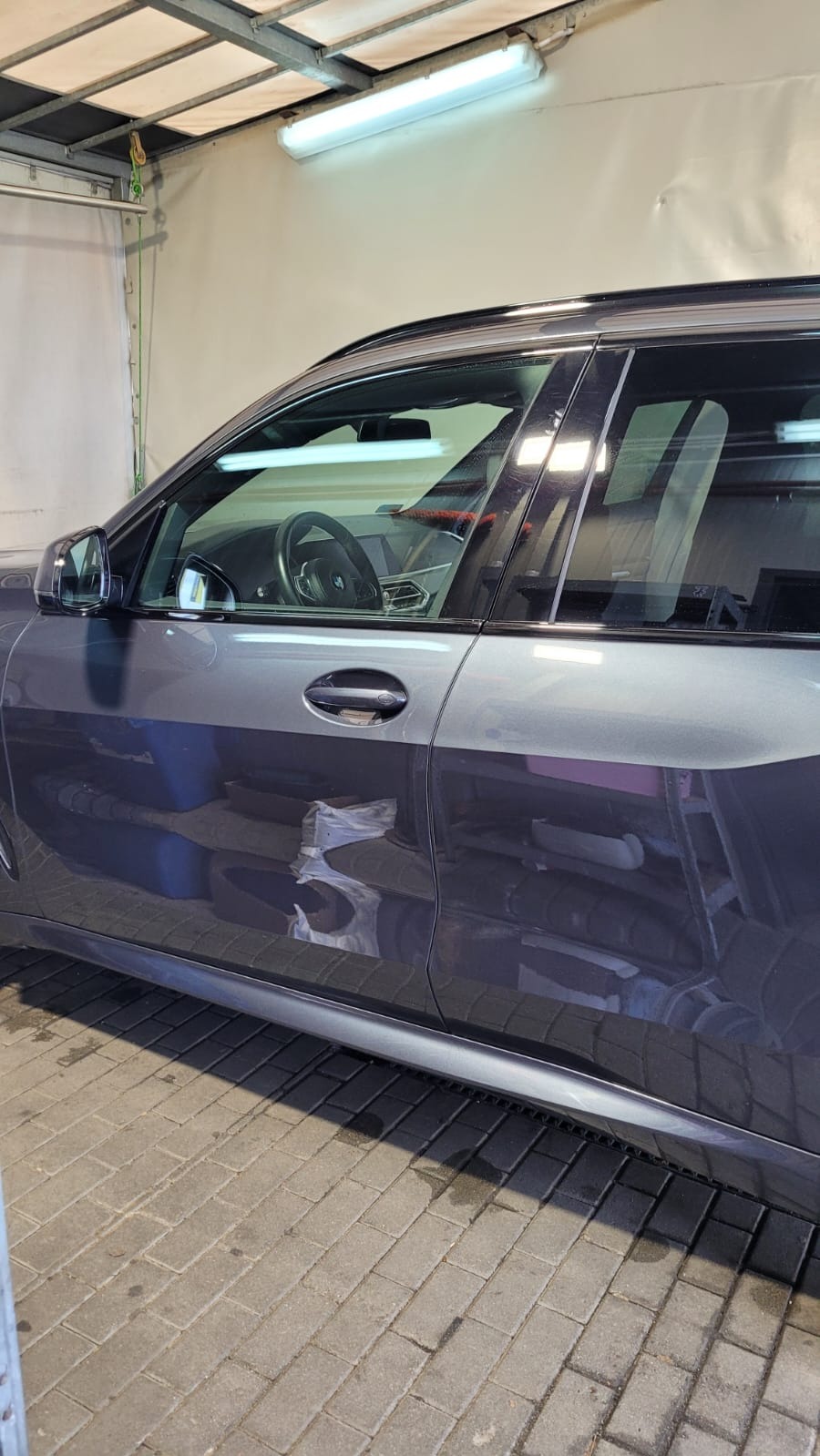 Szary SUV marki BMW, częściowo umyty, z widocznymi śladami wody na karoserii, zaparkowany w myjni z szarymi płytkami na podłodze i oświetleniem jarzeniowym.