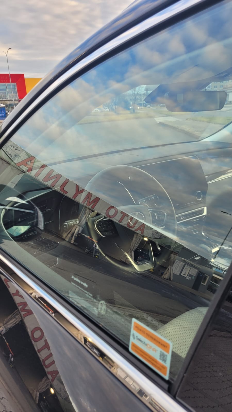 Wnętrze samochodu widoczne przez szybę z naklejką i odbiciem myjni samochodowej.