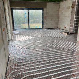 HYDRO SOLUTIONS SEBASTIAN SZYMAŃSKI - Białe rury ogrzewania podłogowego ułożone na folii z czerwoną siatką w surowym pomieszczeniu z oknem, widoczne elementy instalacji elektrycznej na ścianie.