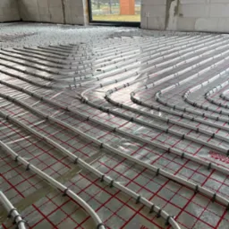 Rozłożone rury ogrzewania podłogowego na folii refleksyjnej z czerwonym nadrukiem kratki, widok z góry, w surowym stanie, ściany z białego betonu komórkowego.