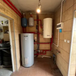 Kotłownia z dwoma zbiornikami Ferroli, instalacją z czerwonych rur, zabezpieczeniami i rozdzielnicą elektryczną na beżowych płytkach, oświetlona wiszącą żarówką.