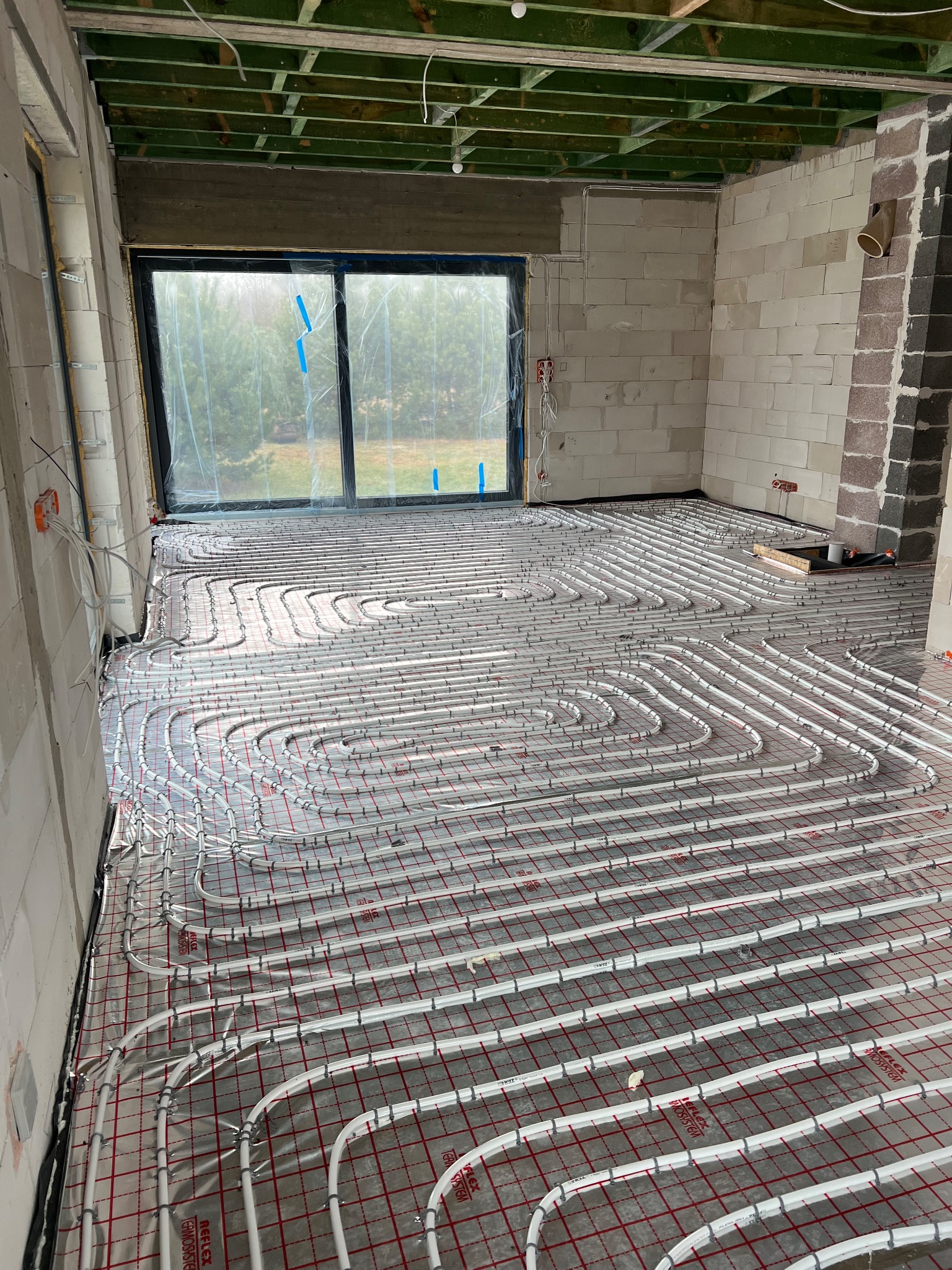 Białe rury ogrzewania podłogowego ułożone na folii z czerwoną siatką w surowym pomieszczeniu z oknem, widoczne elementy instalacji elektrycznej na ścianie.