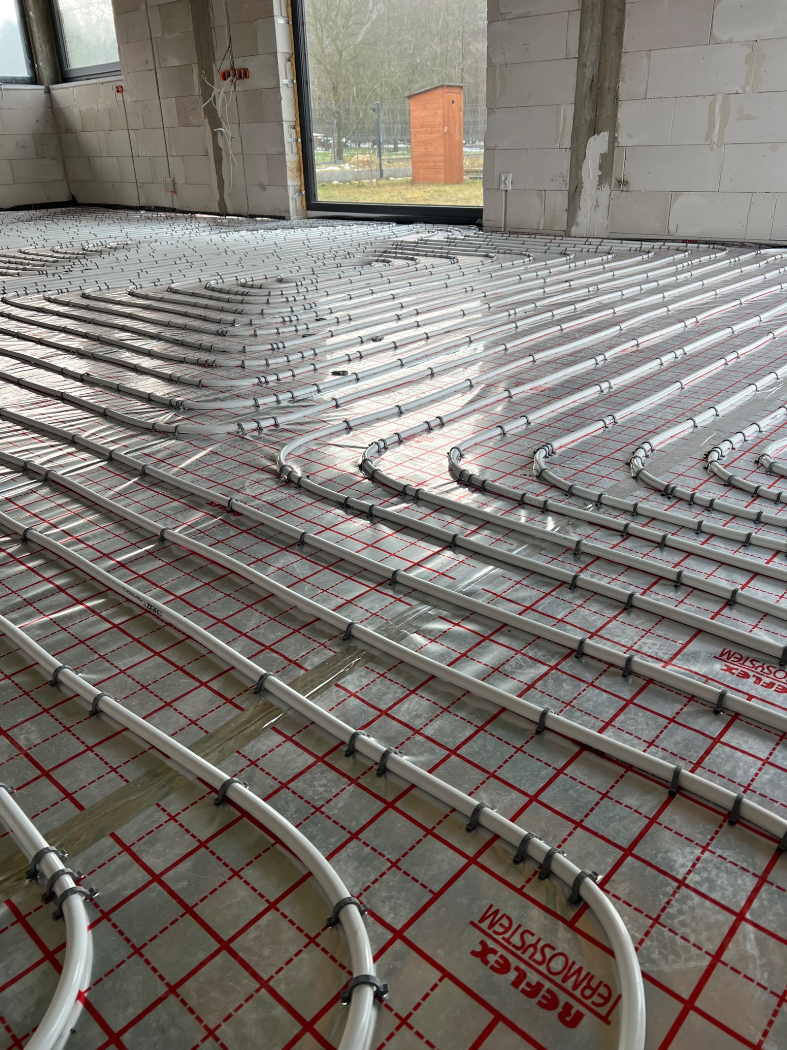 Rozłożone rury ogrzewania podłogowego na folii refleksyjnej z czerwonym nadrukiem kratki, widok z góry, w surowym stanie, ściany z białego betonu komórkowego.
