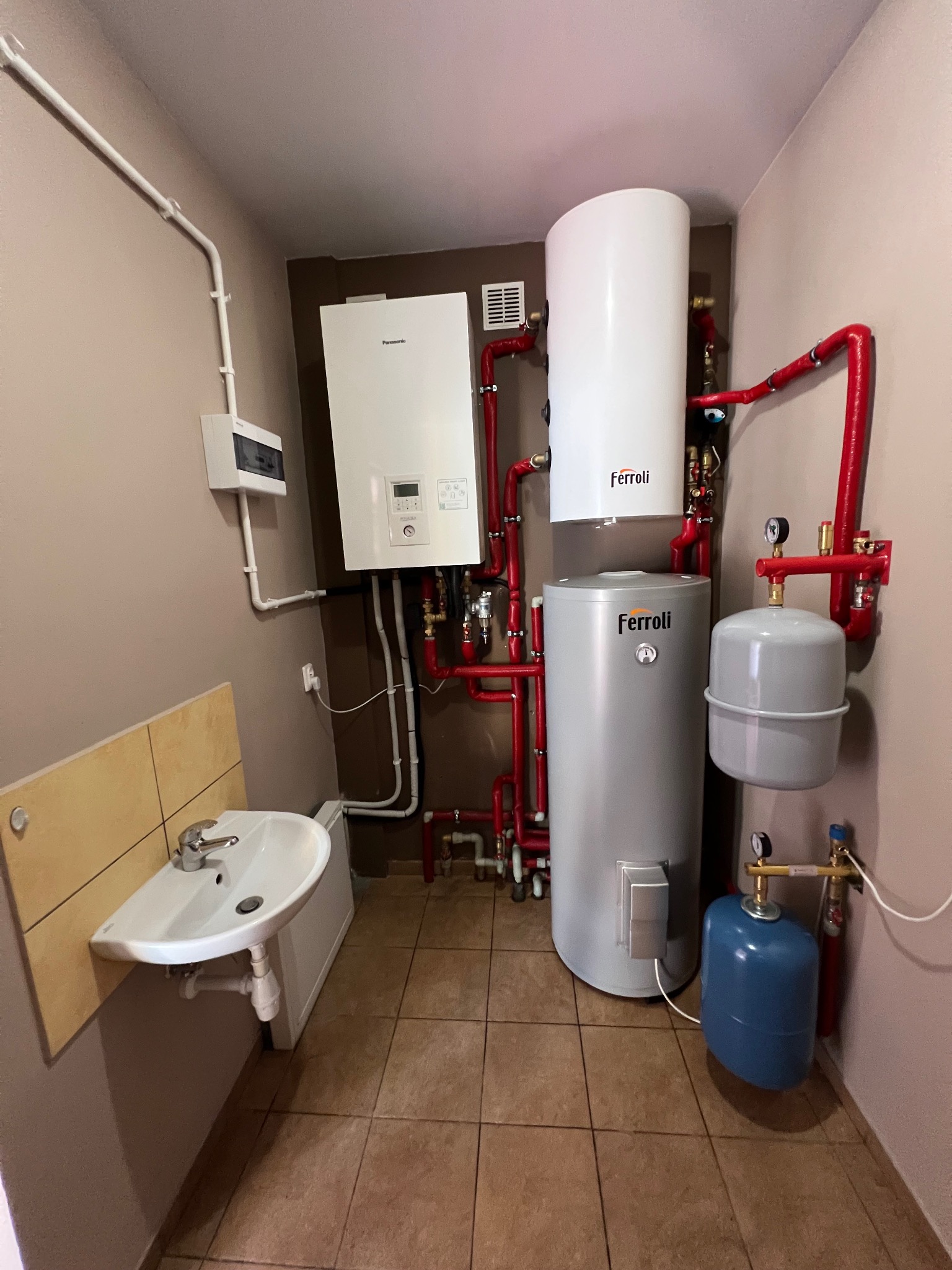 Kotłownia z piecem gazowym Panasonic, zbiornikiem Ferroli, oraz rozbudowaną instalacją z czerwonych rur i elementami hydraulicznymi. Obok umywalka z szafką i płytkami na ścianie.